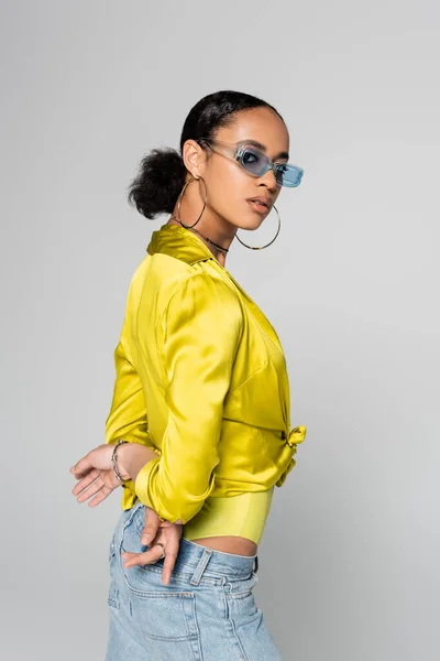 young african american model in blue sunglasses and trendy clothes posing with hands behind back isolated on grey