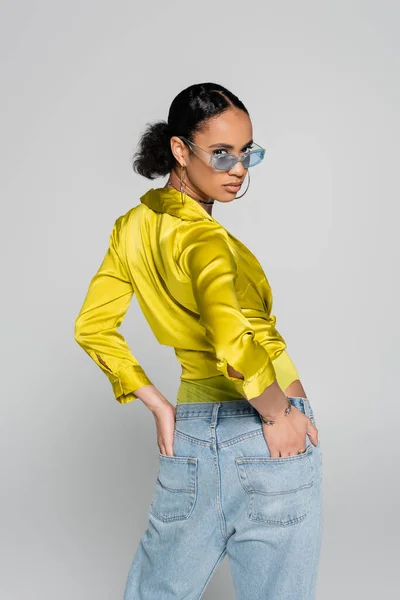 年轻的非洲模特 戴着蓝色太阳镜 穿着时髦的衣服 手插在后面的口袋里 与灰色隔离 — 图库照片