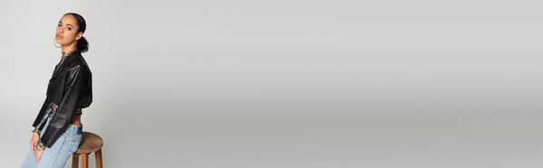 довольно африканская американка в обрезанной кожаной куртке и джинсах опираясь на деревянный стул на сером фоне, баннер 