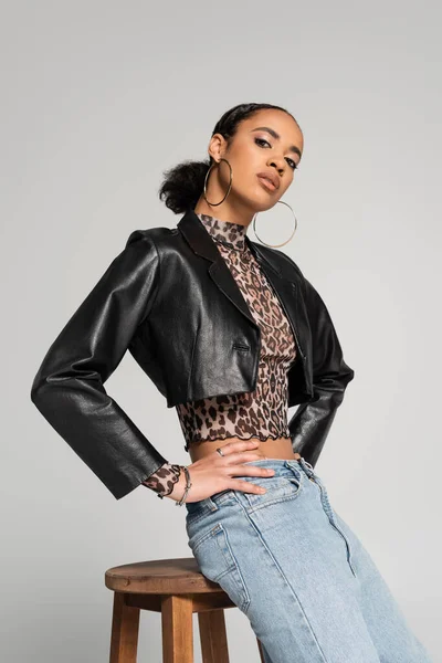 stylish african american model in cropped jacket and jeans posing with hands on hips near wooden high chair isolated on grey