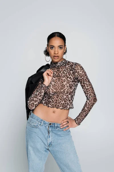 fashionable african american woman in cropped top with animal print holding black jacket while posing isolated on grey