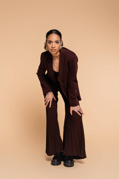 full length of young african american woman in stylish blazer with wide pants posing on beige 