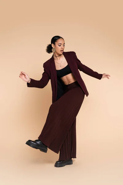 stock image full length of african american woman in trendy maroon suit with wide leg pants posing on beige 