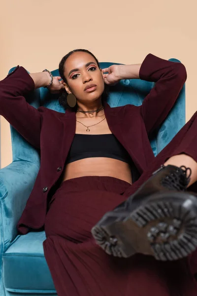 stock image young african american model in stylish suit showing black boot while sitting on blue velvet armchair isolated on beige 