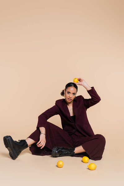 young african american woman in stylish burgundy suit sitting around fresh lemons on beige 