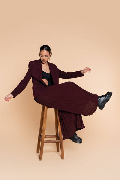 full length of young african american model in burgundy suit sitting on wooden high stool on beige 
