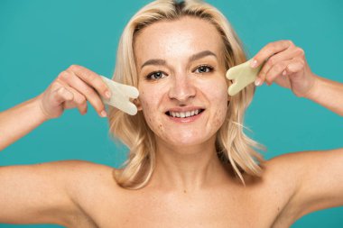 Yüzünde lekeler olan neşeli bir kadın. Yüzüne masaj yapan, turkuazdan izole edilmiş yeşim yüzlü bir kazıyıcı.