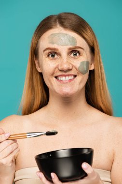 Turkuazda izole edilmiş kilden maske takan kızıl saçlı, elinde kâse ve kozmetik fırçayla mutlu bir kadın. 