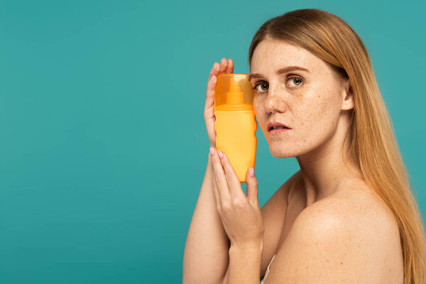 Freckled woman holding sunscreen and looking at camera isolated on turquoise