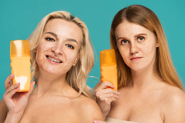 happy women with bare shoulders holding bottles with sunscreen isolated on turquoise 