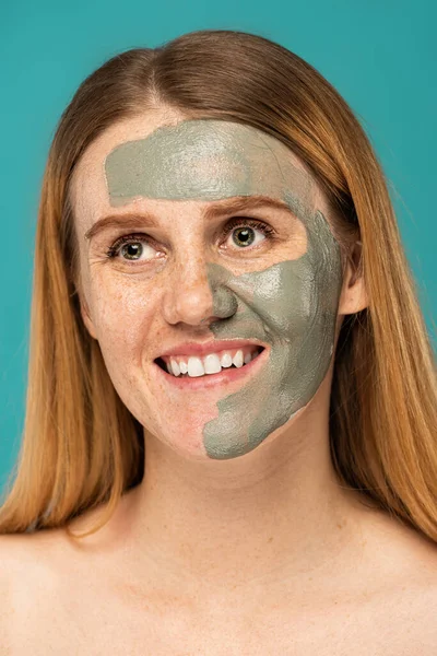 cheerful woman with red hair and clay mask on half of face smiling isolated on turquoise