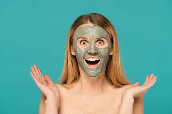amazed woman with red hair and clay mask on face gesturing isolated on turquoise
