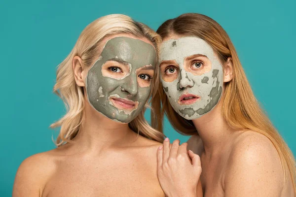 redhead and blonde women with clay mask on faces looking at camera isolated on turquoise