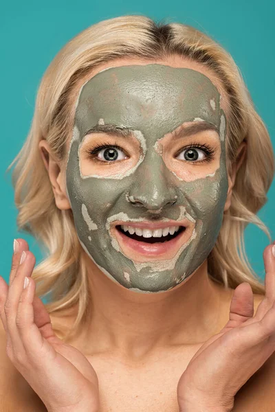excited blonde woman with clay mask on face looking at camera isolated on turquoise