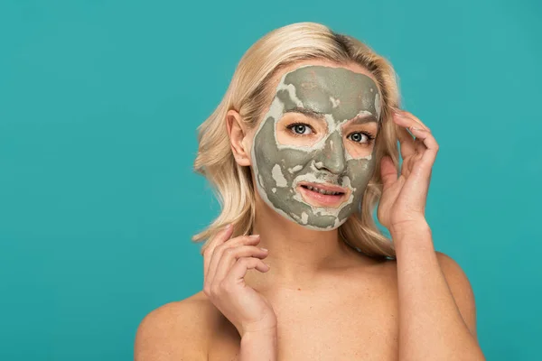blonde woman with clay mask on face looking at camera isolated on turquoise