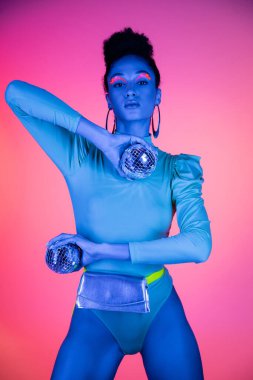 Pembe arka planda disko topları tutan neon yüzlü modaya uygun Afrikalı Amerikalı kadın.