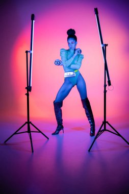 Trendy african american model in bodysuit holding disco balls near neon lamps on purple background clipart