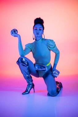 Pembe ve mor arka planda disko topları tutan, deri kıyafetli ve çizmeli Afro-Amerikan model.
