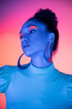 Neon makyajlı genç Afro-Amerikan modeli pembe ve mor arka plana bakıyor.