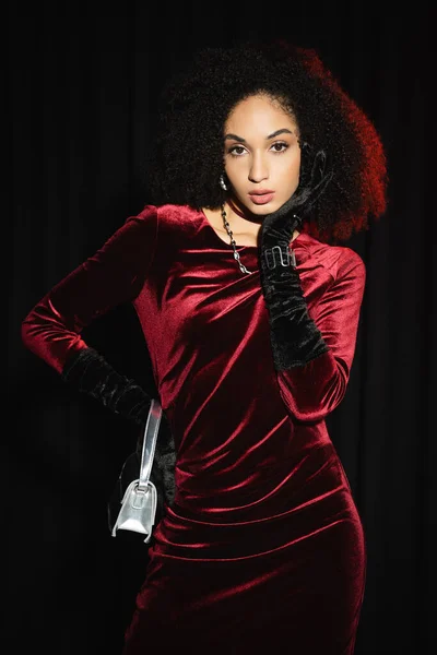 Trendy african american model in velvet dress and gloves looking at camera isolated on black