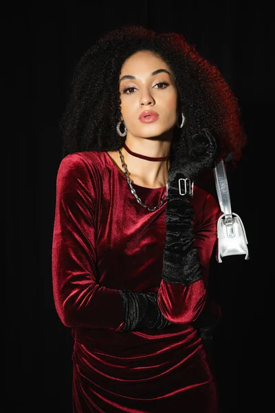 Fashionable african american model in burgundy dress and handbag isolated on black