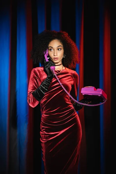Stylish african american model in velvet dress talking on telephone near background with blue light