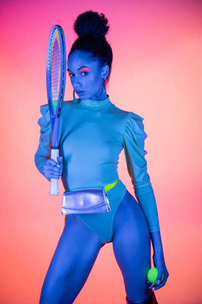 Модная африканская американка в боди-костюме держит теннисную ракетку и мяч на розовом фоне с неоновым светом 