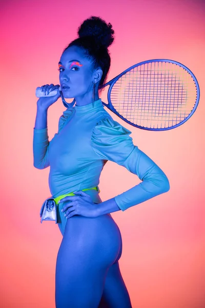 stock image Trendy african american woman in bodysuit holding tennis racket on pink background with neon light 