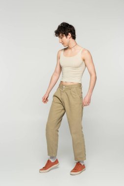 full length of young pansexual person in beige pants and sneakers standing on grey background clipart