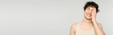 cheerful pansexual model in tank top covering face with hand and smiling with closed eyes isolated on grey, banner clipart