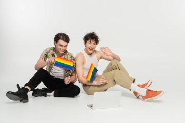 LGBT bayrağı taşıyan mutlu çiftler gri arka planda otururken bilgisayarlarında görüntülü sohbet ediyorlar.