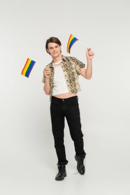 Siyah pantolonlu ve yılan derisi bluzlu tam boy panseksüel model gri arka planda küçük lgbt bayrakları taşıyor.