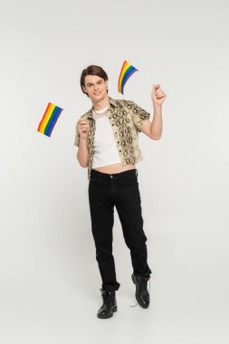 full length of positive pansexual model in black pants and snakeskin print blouse holding small lgbt flags on grey background clipart