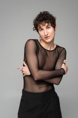 Siyah şeffaf üstlü esmer panseksüel model. Gri üzerinde izole edilmiş çapraz kollarla poz veriyor.