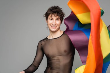 Siyah şeffaf üstlü neşeli olmayan kişi ve gri üzerinde lgbt bayrağı tutan gümüş aksesuarlar.