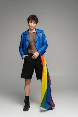 LGBT bayrağı taşıyan, mavi deri ceketli ve gri arka planda siyah şortlu tam boy panseksüel insan.