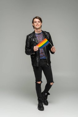 full length of carefree pangender person in black ripped pants and leather jacket standing with small lgbt flag on grey background clipart