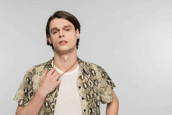 young nonbinary person in snakeskin print blouse touching golden necklace and looking at camera isolated on grey