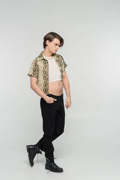 full length of pansexual person in snakeskin print blouse and black trousers posing with hand in pocket on grey background