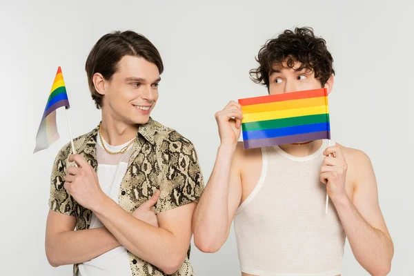 young nonbinary person obscuring face with small lgbt flag near smiling friend isolated on grey