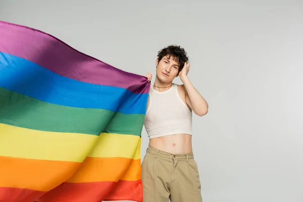stylish bigender person in crop top and beige pants standing near rainbow flag isolated on grey