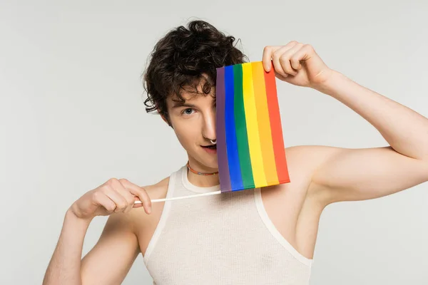 young nonbinary person obscuring face with small lgbt flag and looking at camera isolated on grey