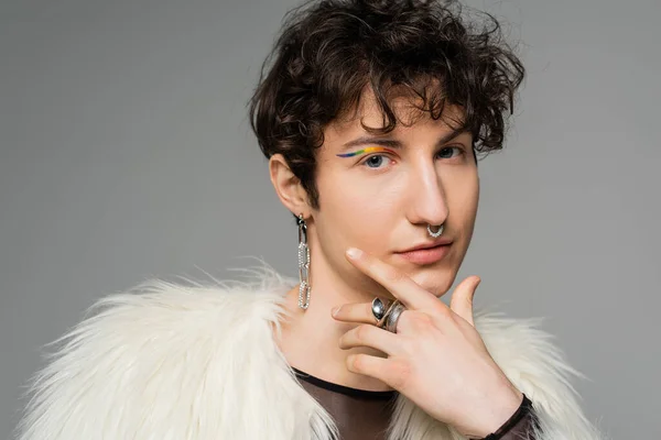 stock image portrait of brunette nonbinary person with rainbow eye liner and silver accessories holding hand near face isolated on grey
