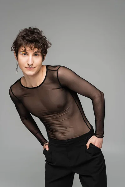 trendy bigender person in black transparent top posing with hands in pockets isolated on grey