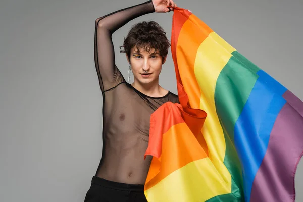 brunette bigender person in black transparent top holding rainbow flag isolated on grey