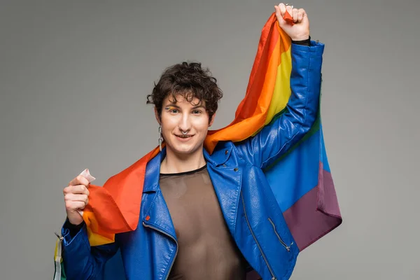 穿着蓝色皮夹克的无忧无虑的泛性别者 手持彩虹旗 凝视着用灰色隔离的相机 — 图库照片