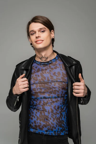 joyful pansexual model in animal print top and black leather jacket smiling at camera isolated on grey