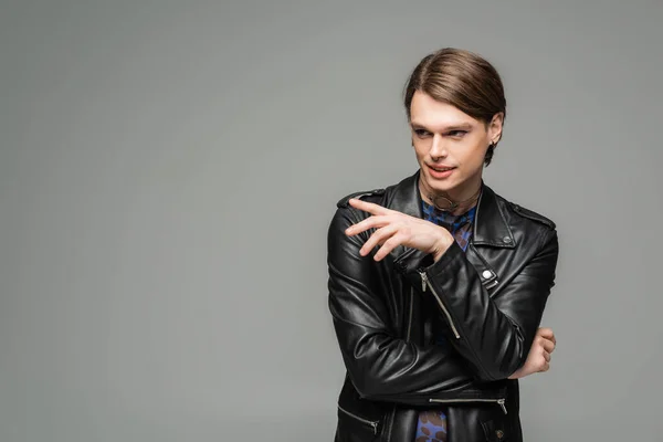 fashionable bigender person in black leather jacket smiling and pointing with finger isolated on grey