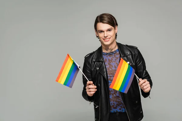 happy pangender person in black leather jacket holding small lgbt flags and smiling at camera isolated on grey