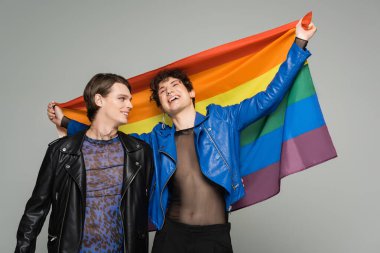 Mavi deri ceketli kaygısız çift cinsiyetli kişi gri renkte izole edilmiş moda partneri yanında gökkuşağı bayrağı tutuyor.